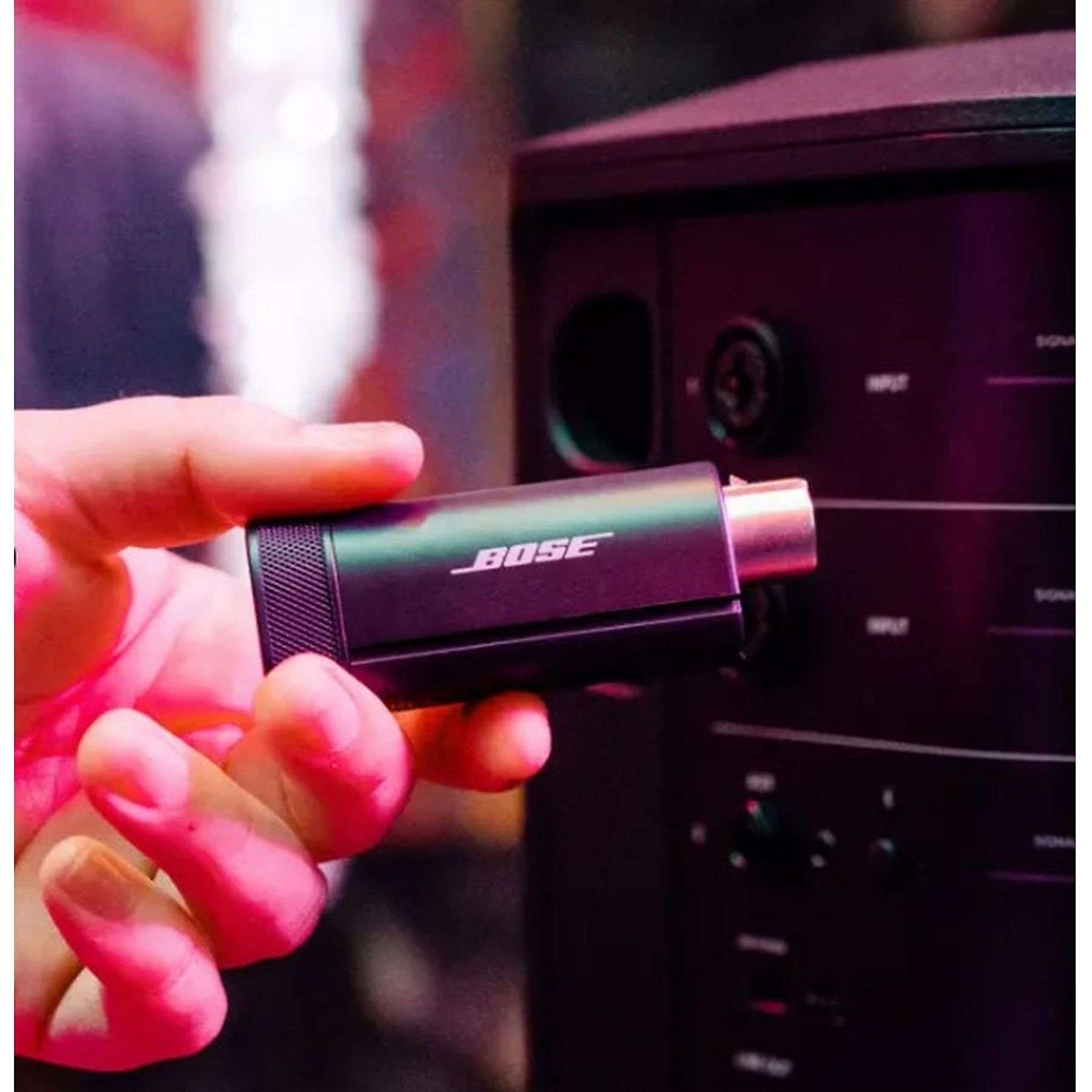Ręka trzyma czarny adapter Bose ze srebrnym złączem, w pobliżu czarnego urządzenia audio. Tło jest rozmyte.