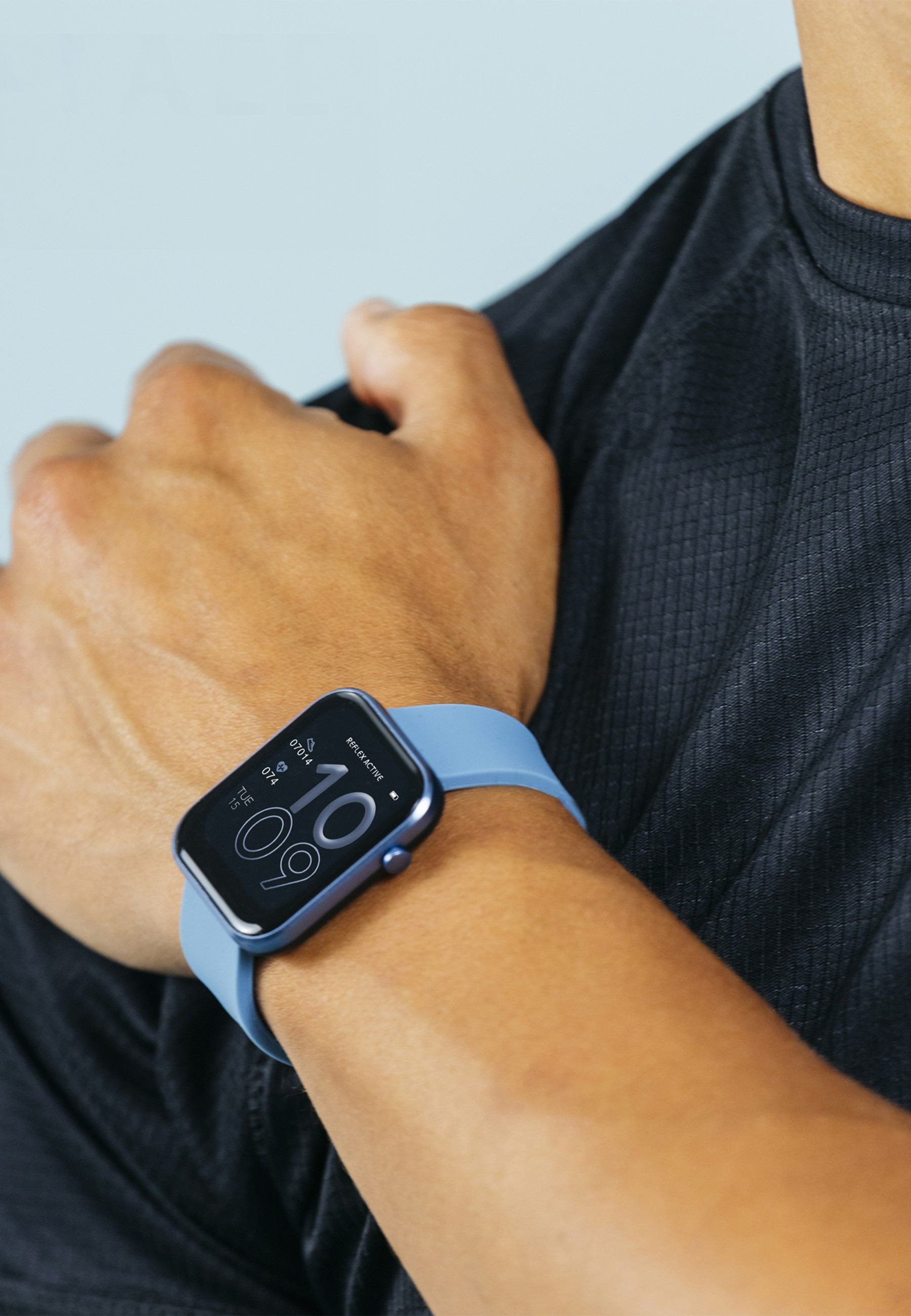 Eine Person, die eine blaue Smartwatch mit einem schwarzen Hemd trägt. Die Uhr zeigt die Zeit an. Der Hintergrund ist hellblau.