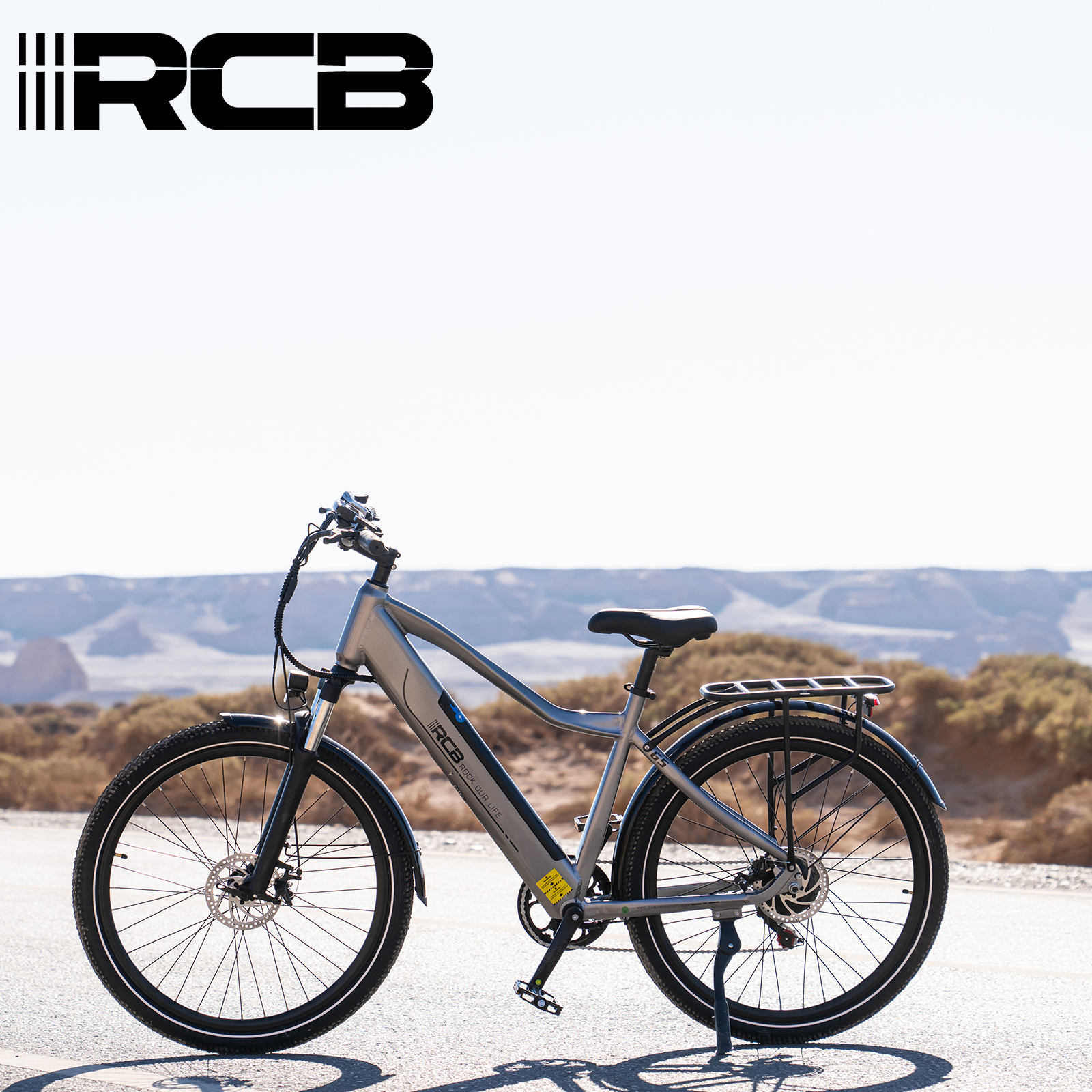 Graues Elektrofahrrad auf einer Straße mit Wüstenlandschaft im Hintergrund unter blauem Himmel.