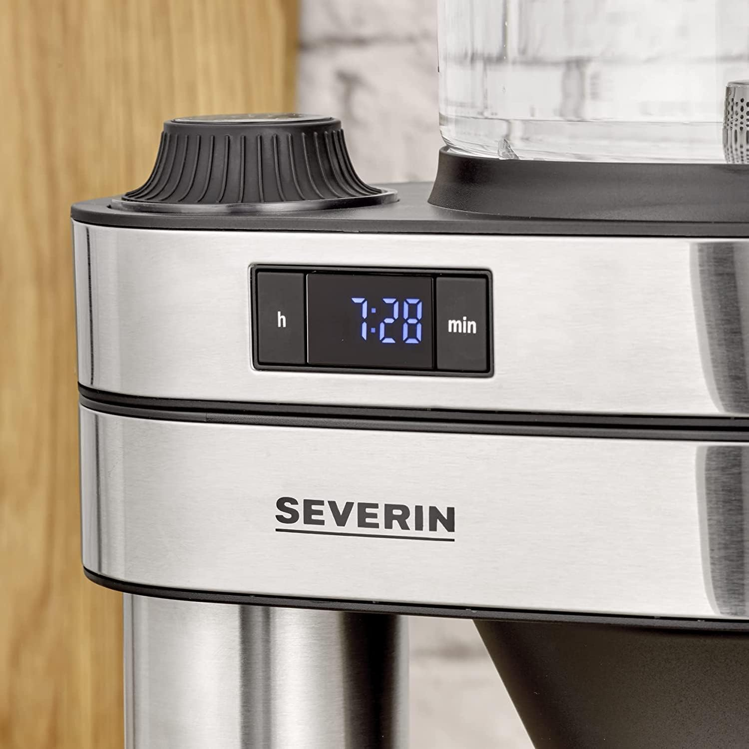 Close-up of a Severin coffee maker. Display shows 7:28. Black details on metallic body. Wooden and brick background.