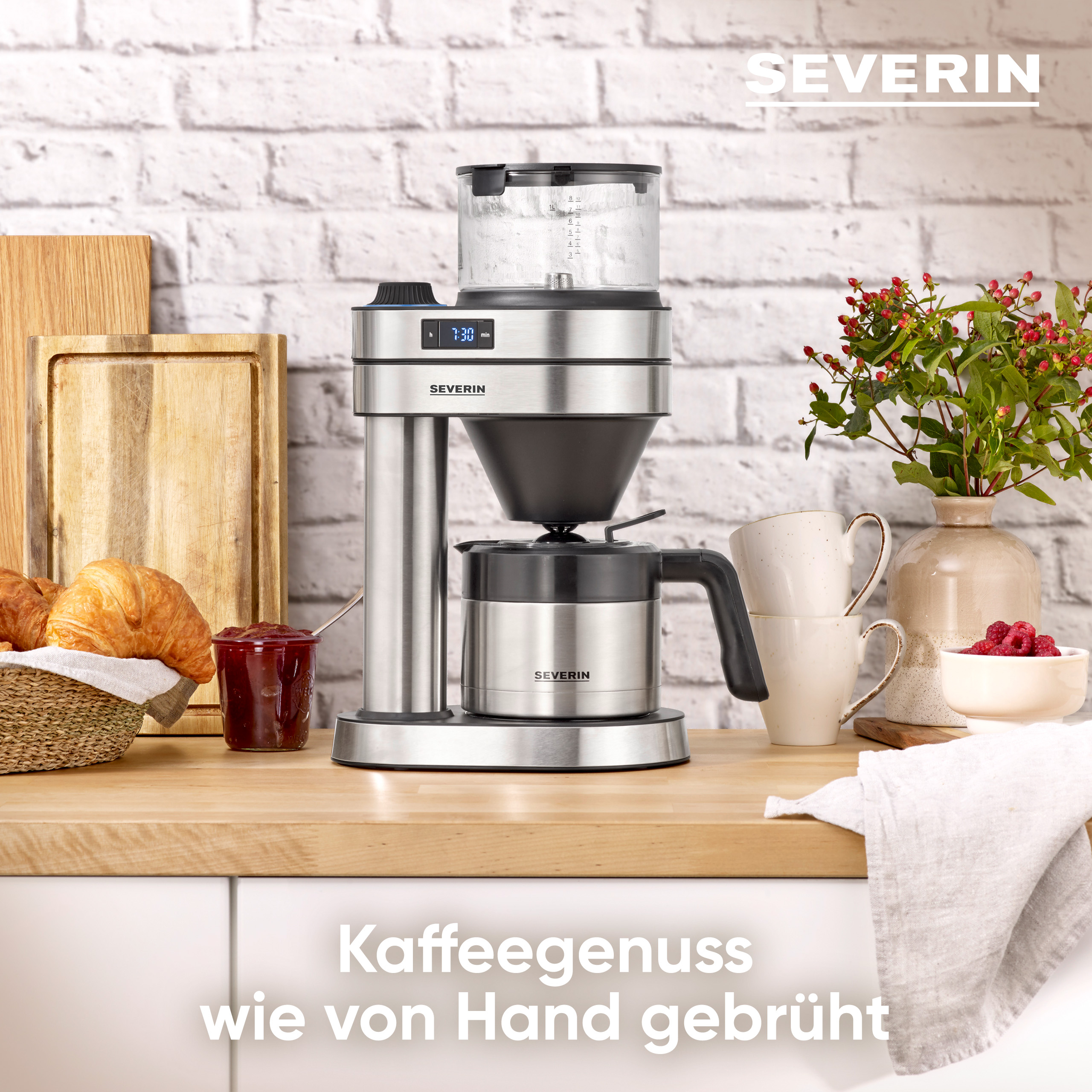 A coffee machine, croissants, and jam are on a wooden counter. A vase of flowers and cups are also present.