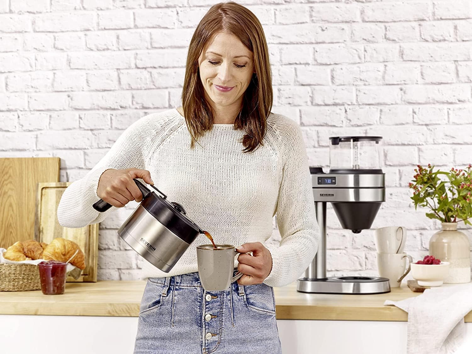 A woman pours coffee from a silver pot into a mug. A coffee maker, croissants, and a vase of flowers are visible.
