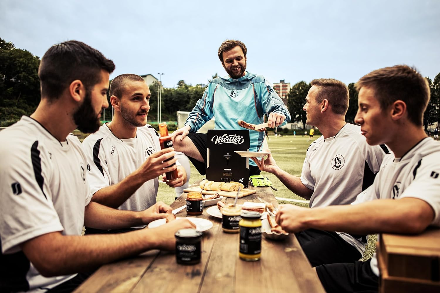 Men in soccer uniforms around a table with food, a man grilling sausages, and a soccer field in the background.