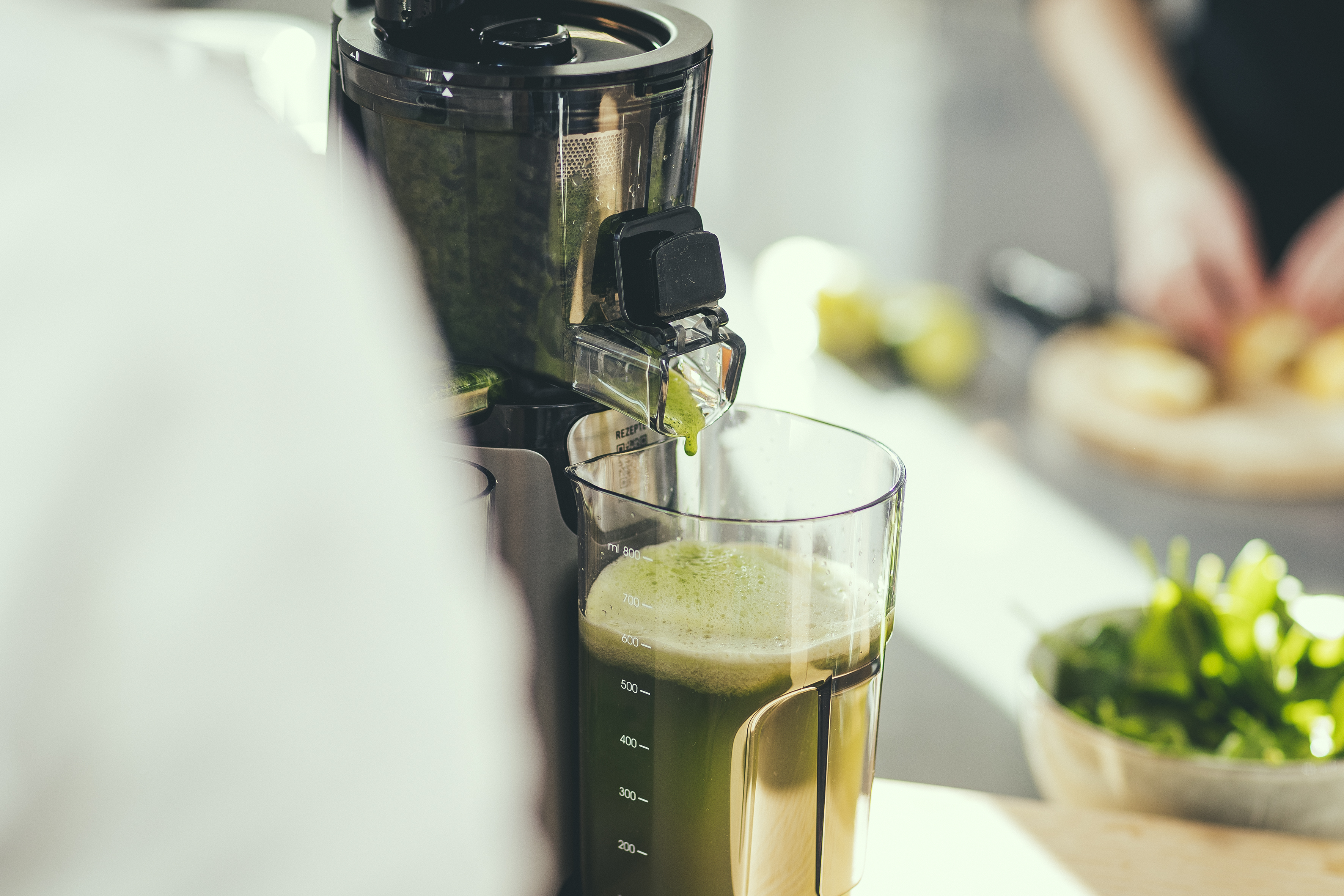 Ein Entsafter gibt grünen Saft in ein Glas ab. Zutaten und eine Person sind im Hintergrund.