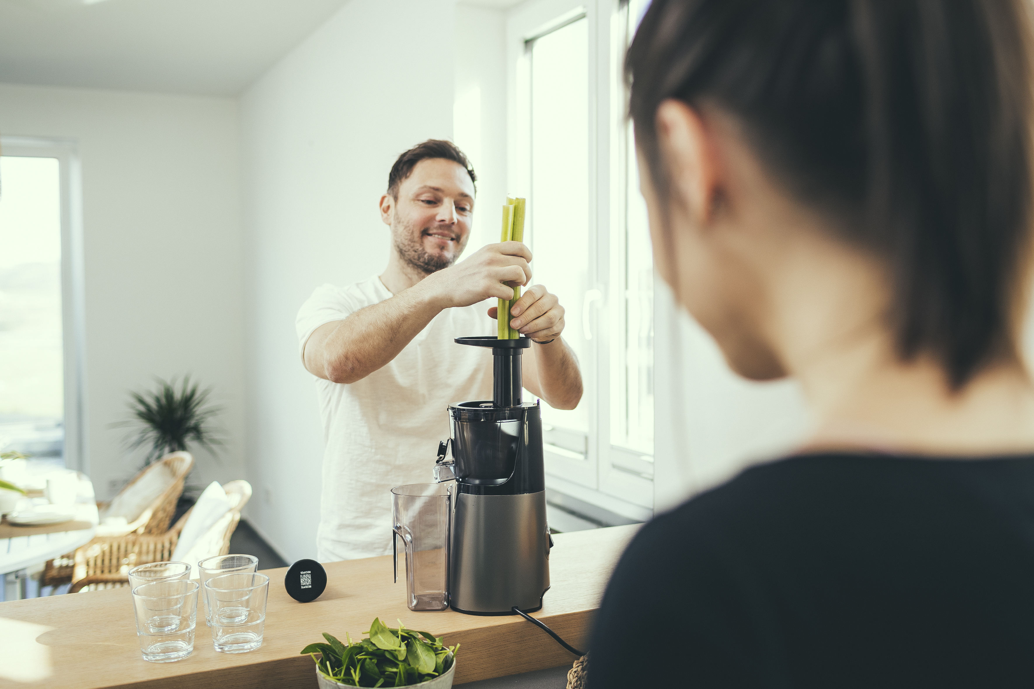 Uomo che estrae il succo di sedano con un'estrattore, un'altra persona è in primo piano. Ambiente cucina con bicchieri e ciotola.