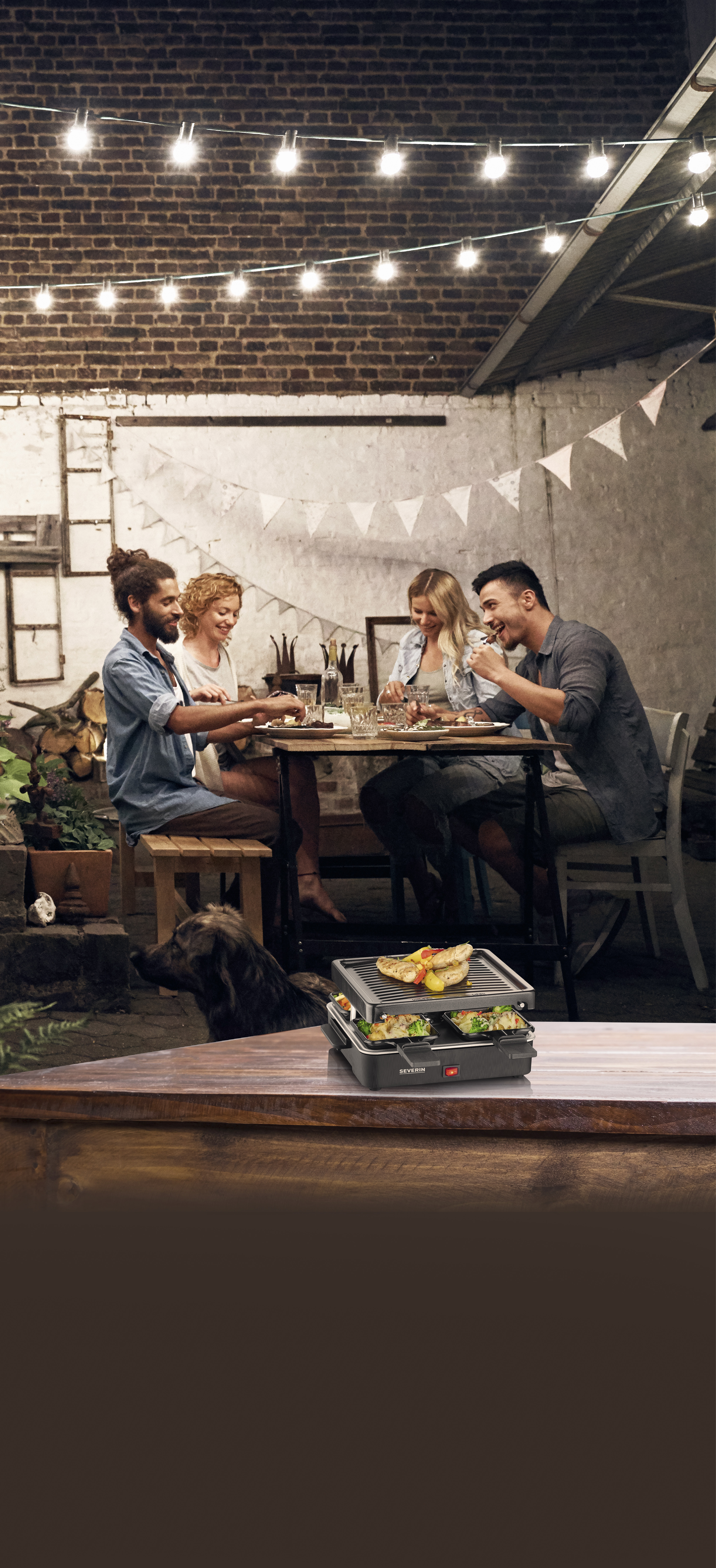 Cztery osoby jedzą na zewnątrz. Grill do raclette jest na stole. Widoczny jest pies. Wiszące światła. Ceglana ściana w tle.