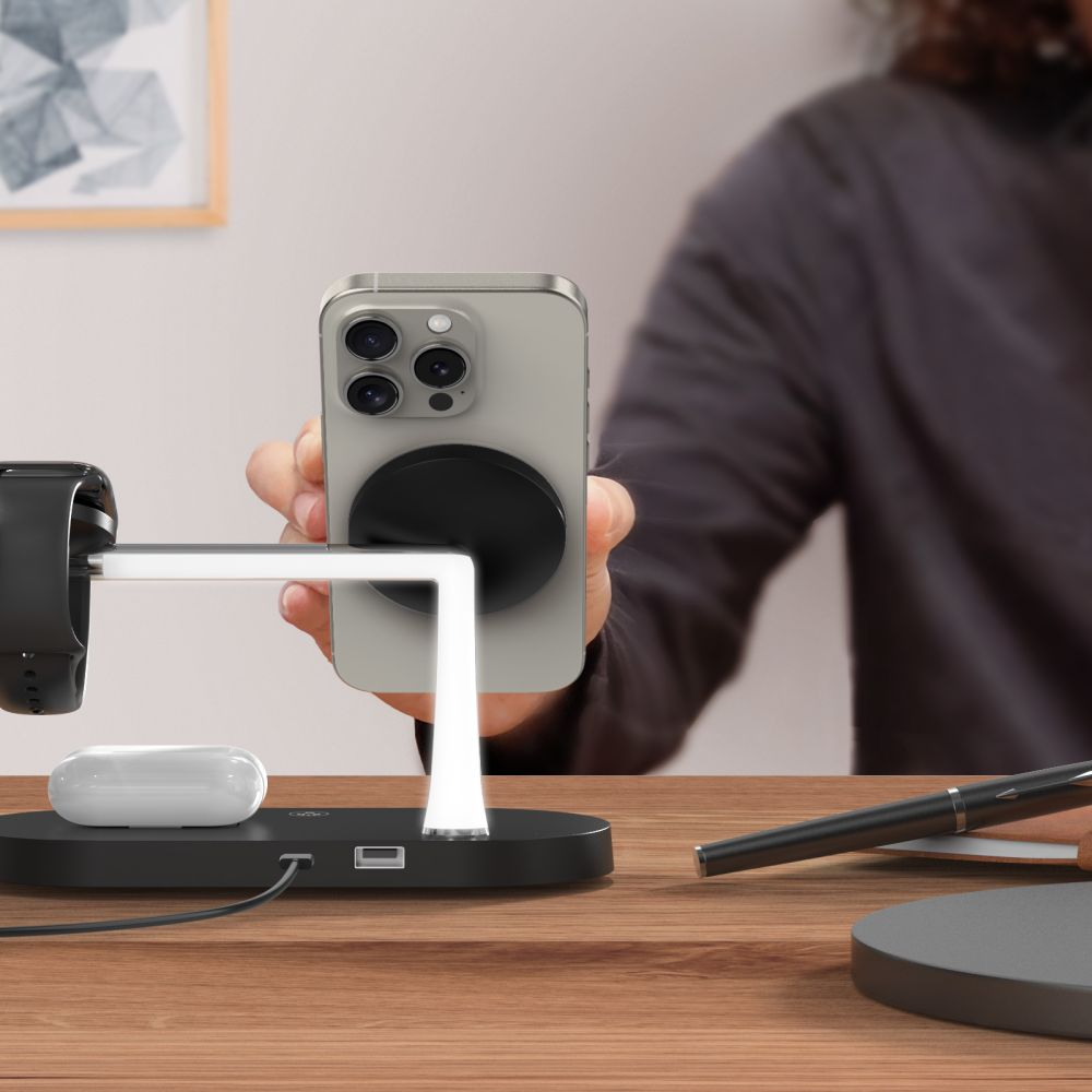 Une personne tient un téléphone sur un chargeur. Le chargeur contient également une Apple Watch, des AirPods et un stylo et un carnet.