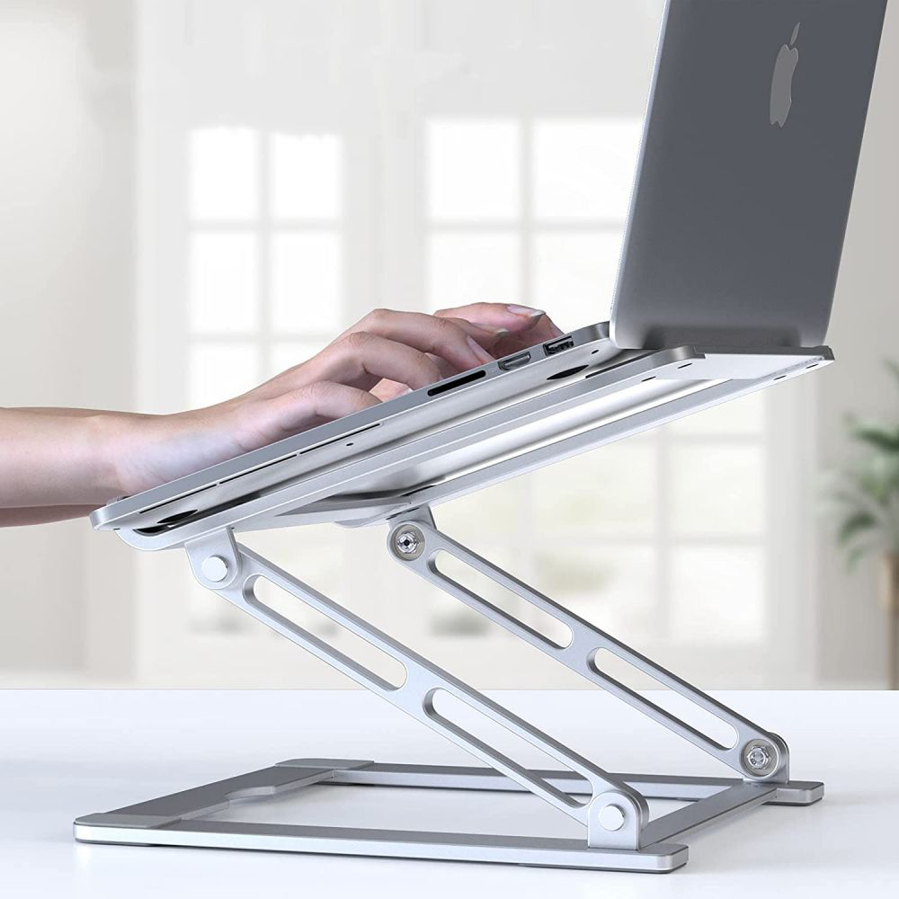 A silver laptop is on a silver adjustable stand. A hand is typing. Background is a white table, window, and plant.