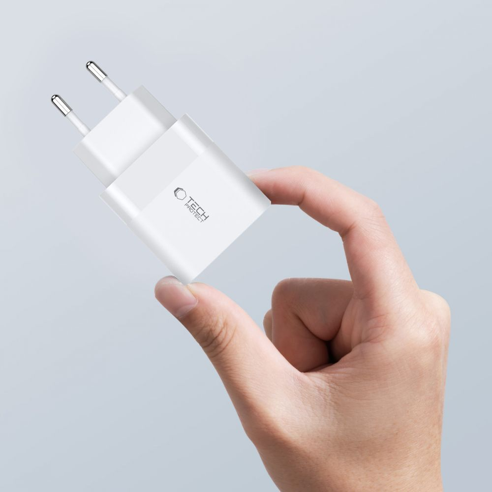 Een hand houdt een witte telefoonlader vast. De lader heeft zilveren pinnen en een 'Tech Protect' logo. De achtergrond is lichtgrijs.