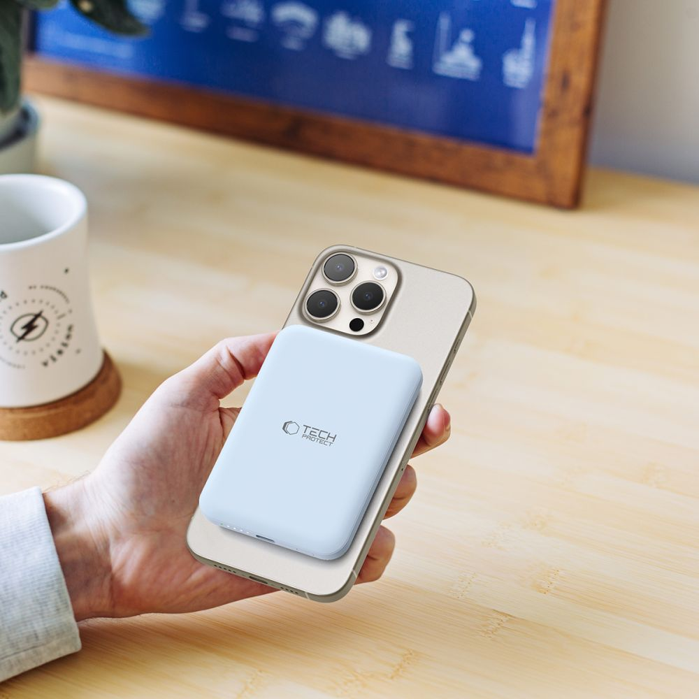 Main tenant un téléphone avec une batterie externe bleu clair attachée. Une tasse blanche et une table en bois sont en arrière-plan.