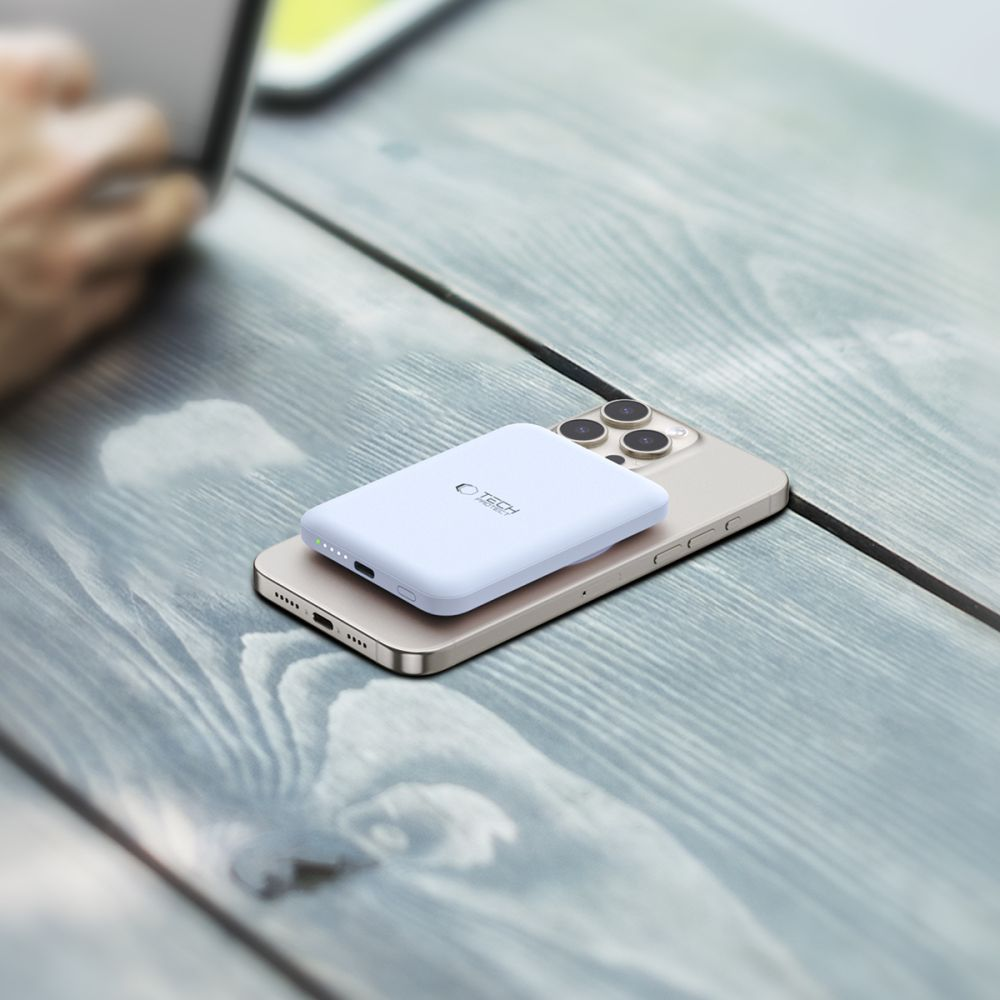Une batterie externe blanche avec un logo est sur un téléphone doré. Les deux sont sur une surface en bois grise. Une main floue est visible.