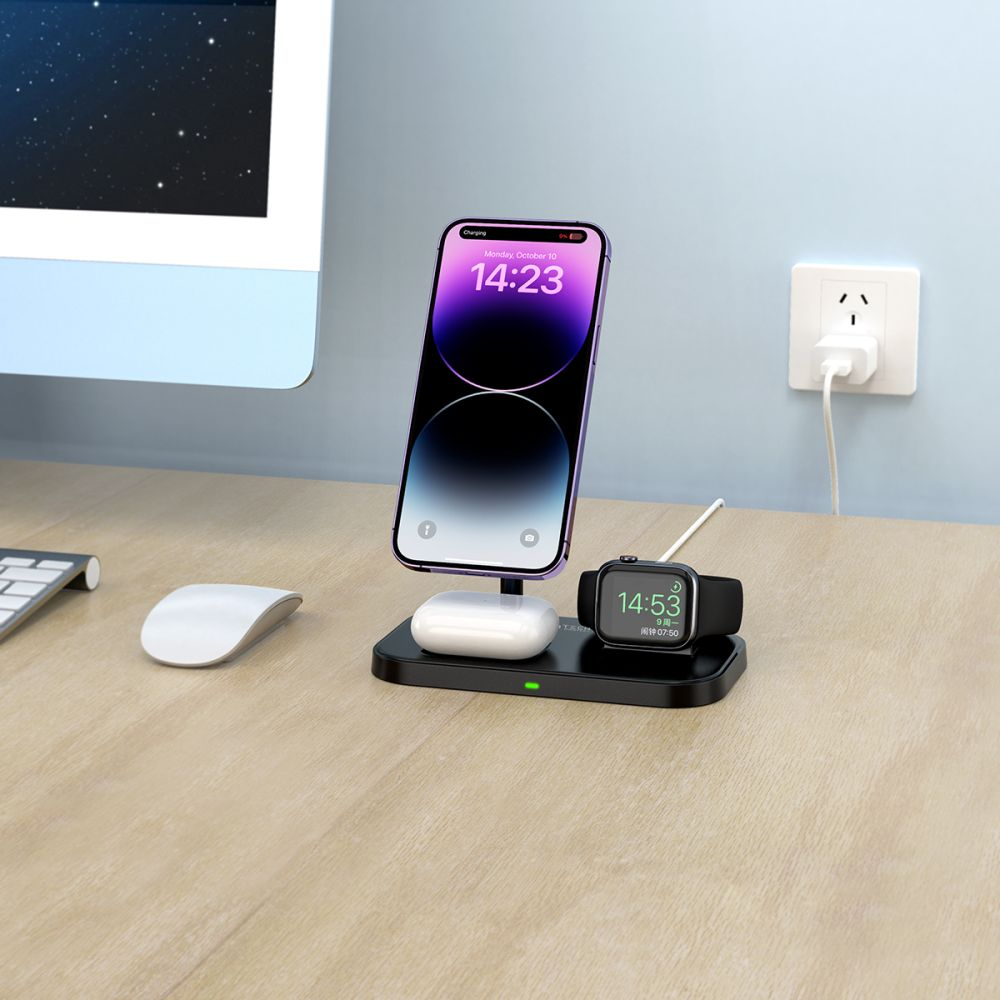 Un teléfono, un reloj y auriculares se cargan en una estación de carga inalámbrica negra. Un monitor de computadora y un ratón son visibles.