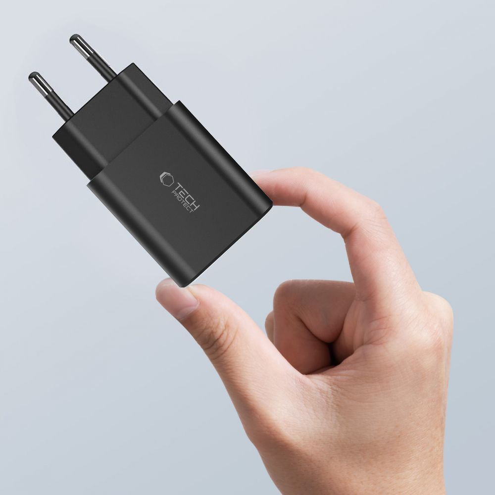 Une main tenant un chargeur Tech Protect noir. Le chargeur a deux broches et un logo. Le fond est gris clair.