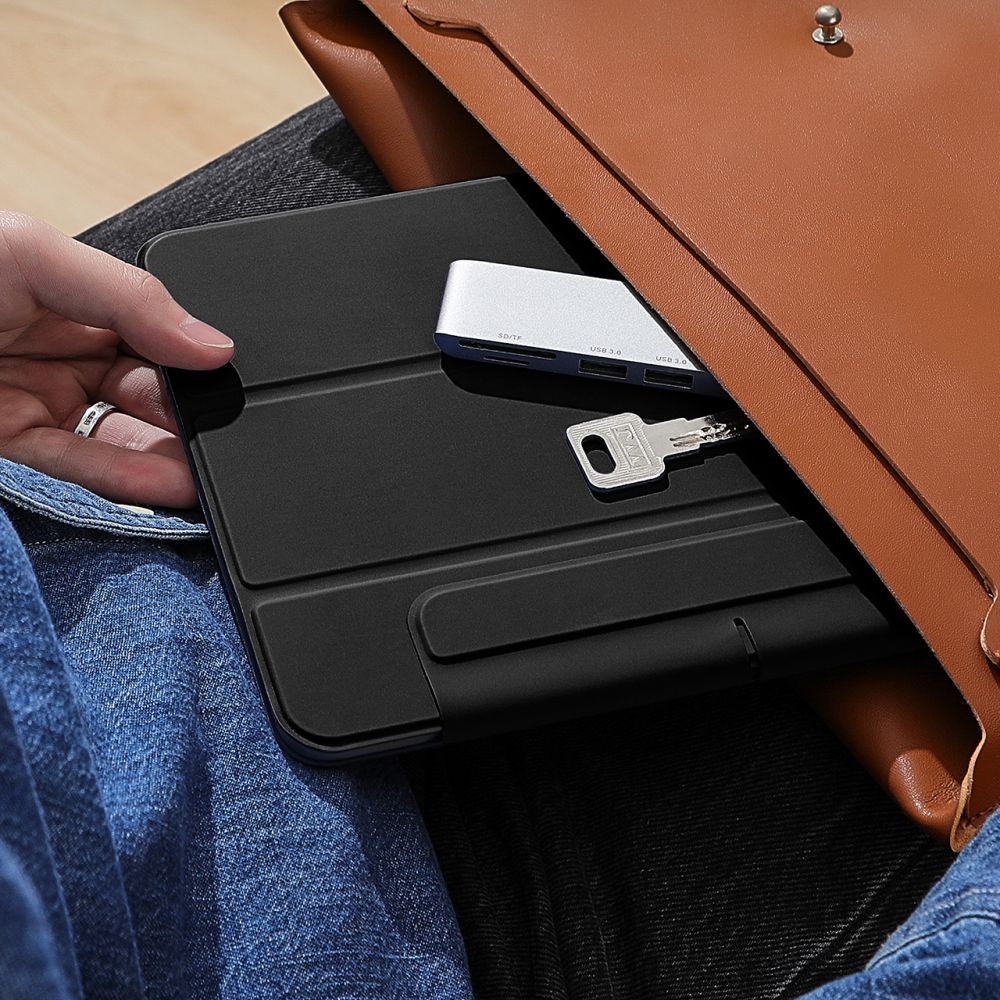 Eine Hand hält eine schwarze Tablethülle. Ein silberner USB-Hub und ein Schlüssel liegen darauf. Im Hintergrund ist eine braune Ledertasche zu sehen.