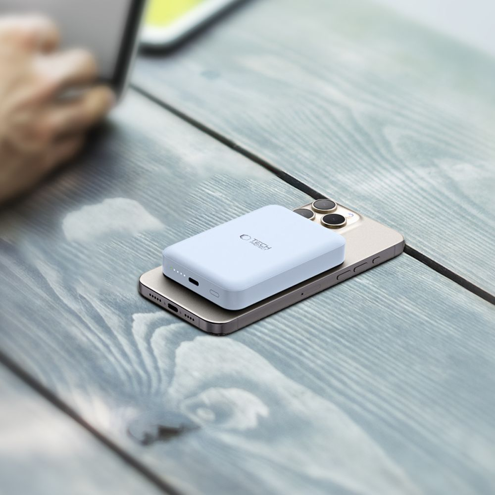 Une batterie externe bleu clair est posée sur un téléphone doré sur une surface en bois grise. Une main floue en arrière-plan utilise un ordinateur portable.