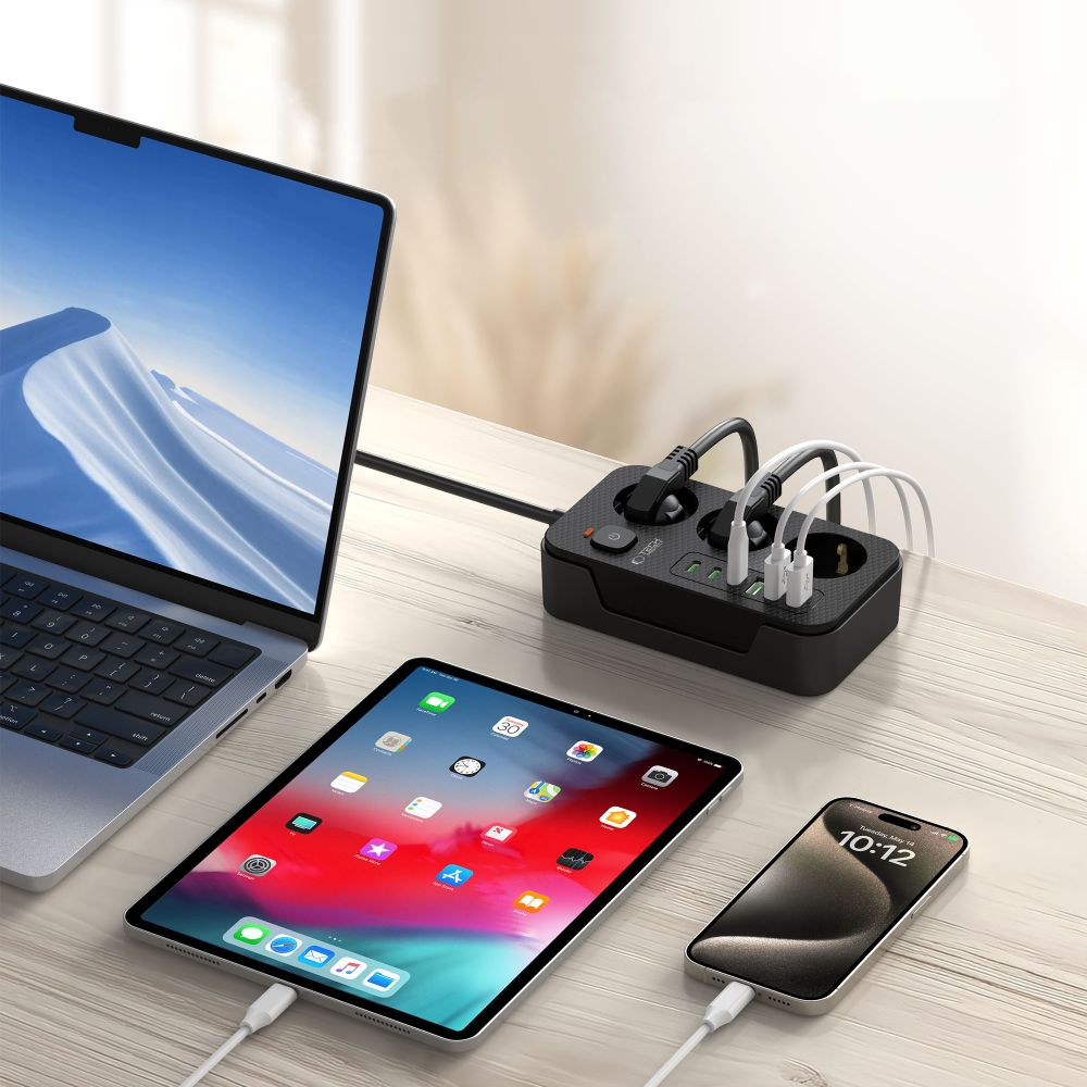 Ordinateur portable, tablette et téléphone en charge sur une table en bois. Une multiprise noire avec plusieurs prises et ports USB est visible.