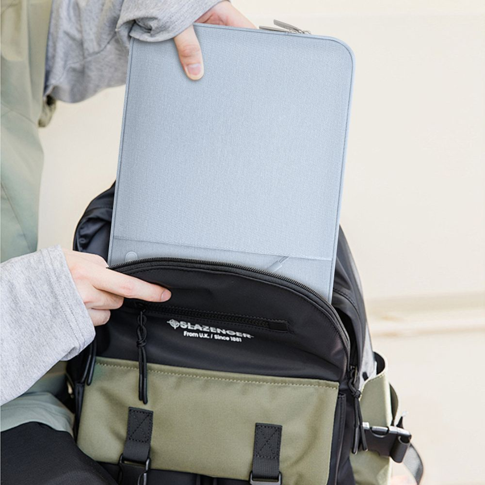 Een persoon plaatst een lichtblauwe laptoptas in een zwarte en olijfgroene rugzak. De rugzak heeft de merknaam Slazenger.