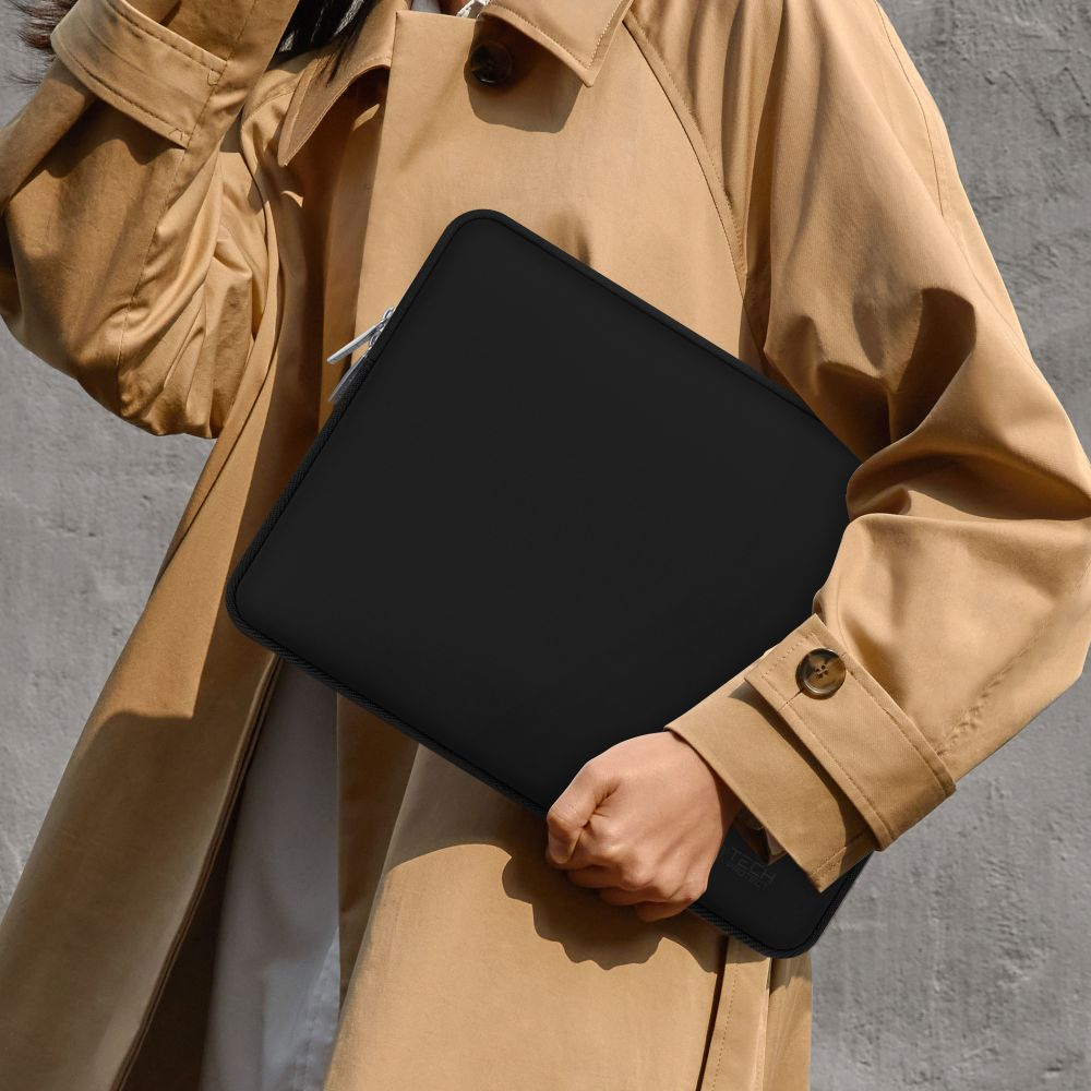 Een persoon in een beige jas houdt een zwarte laptoptas vast. De achtergrond is een grijze muur. De tas heeft een zilveren rits.