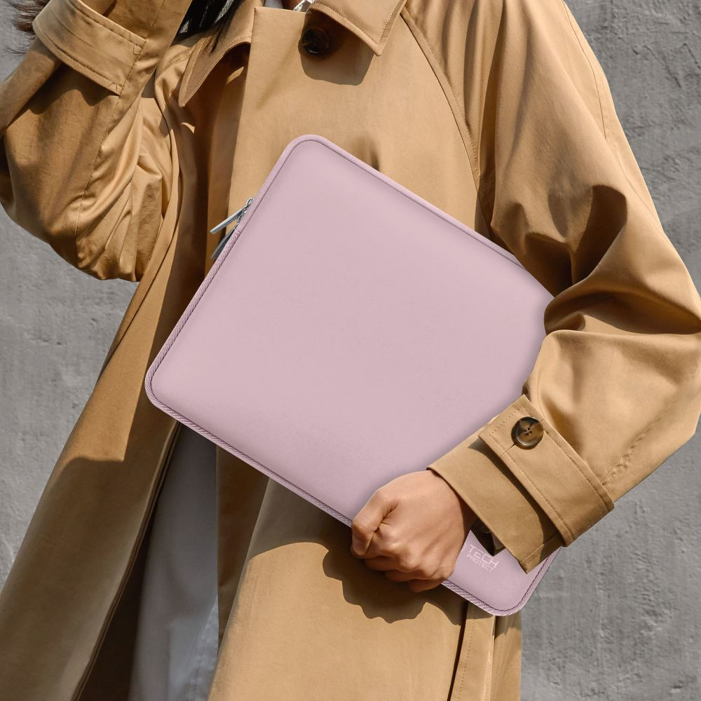 Een persoon houdt een lichtroze laptoptas vast. Ze draagt een beige trenchcoat tegen een grijze muur. De tas heeft een rits.