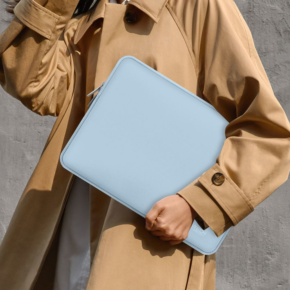 Een persoon houdt een lichtblauwe laptoptas vast. Ze draagt een beige trenchcoat. De achtergrond is een grijze muur.