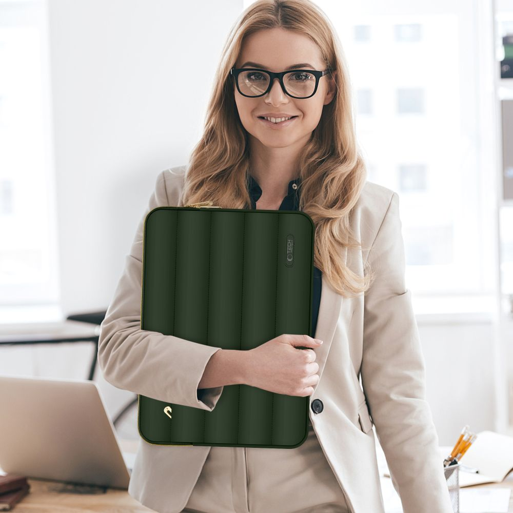 Eine Frau mit Brille hält eine grüne, gepolsterte Laptoptasche. Sie trägt einen beigen Blazer und lächelt. Ein Laptop und Büroutensilien sind im Hintergrund.