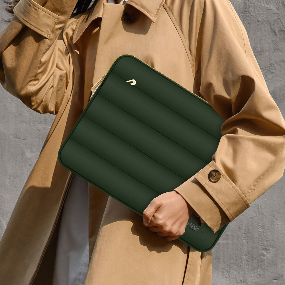 Eine Person hält eine grüne Laptop-Hülle. Die Hülle hat einen goldenen Reißverschluss und ein kleines goldenes Logo. Die Person trägt einen beigen Mantel.