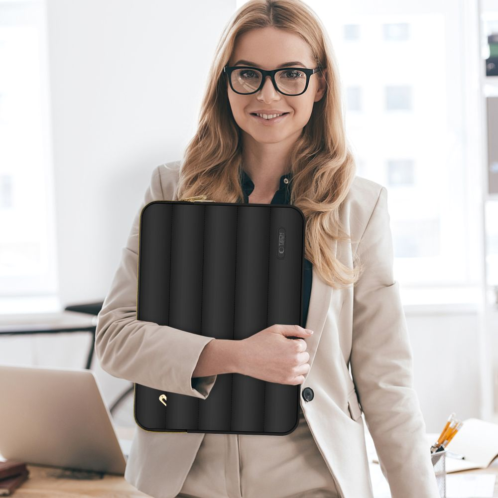 Een vrouw met bril en blazer houdt een zwarte laptoptas vast. Ze glimlacht naar de camera in een helder kantoor.