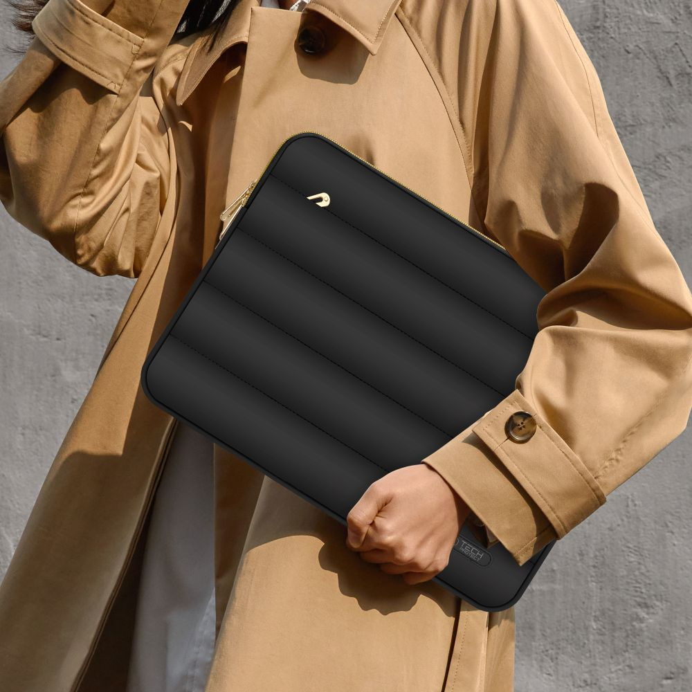 Persoon die een zwarte, gewatteerde laptoptas met een gouden rits vasthoudt. Ze dragen een beige trenchcoat. De achtergrond is een grijze muur.