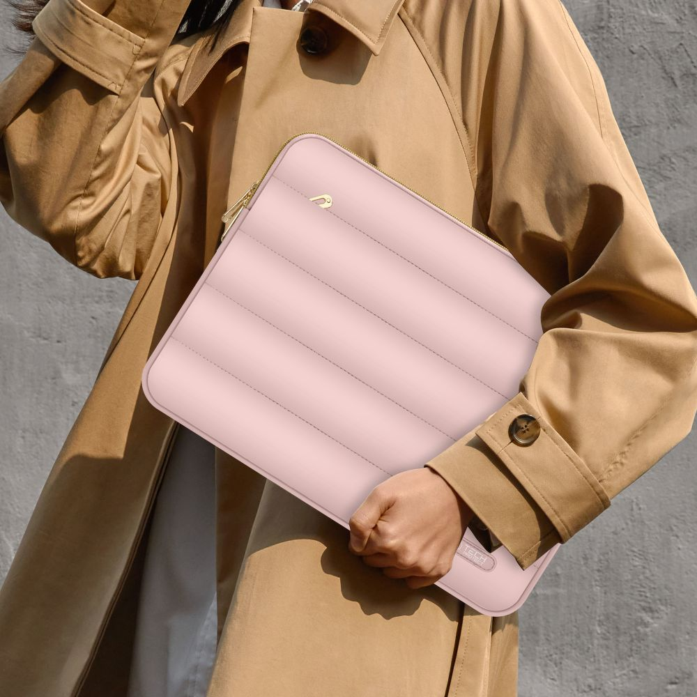 Een persoon houdt een roze gewatteerde laptoptas vast. Ze draagt een beige trenchcoat. De achtergrond is een grijze muur.