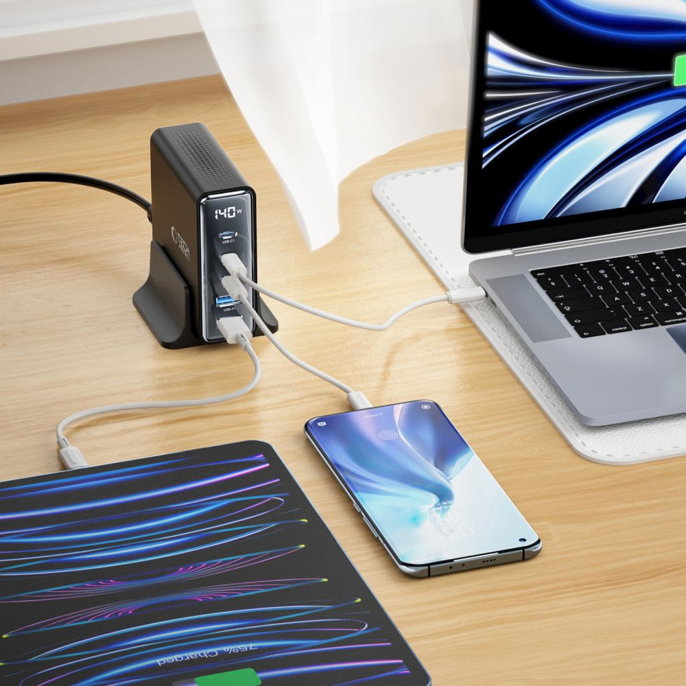 Une station de recharge noire avec des appareils connectés : ordinateur portable, téléphone et tablette. Tous chargent sur un bureau en bois près d'une fenêtre.