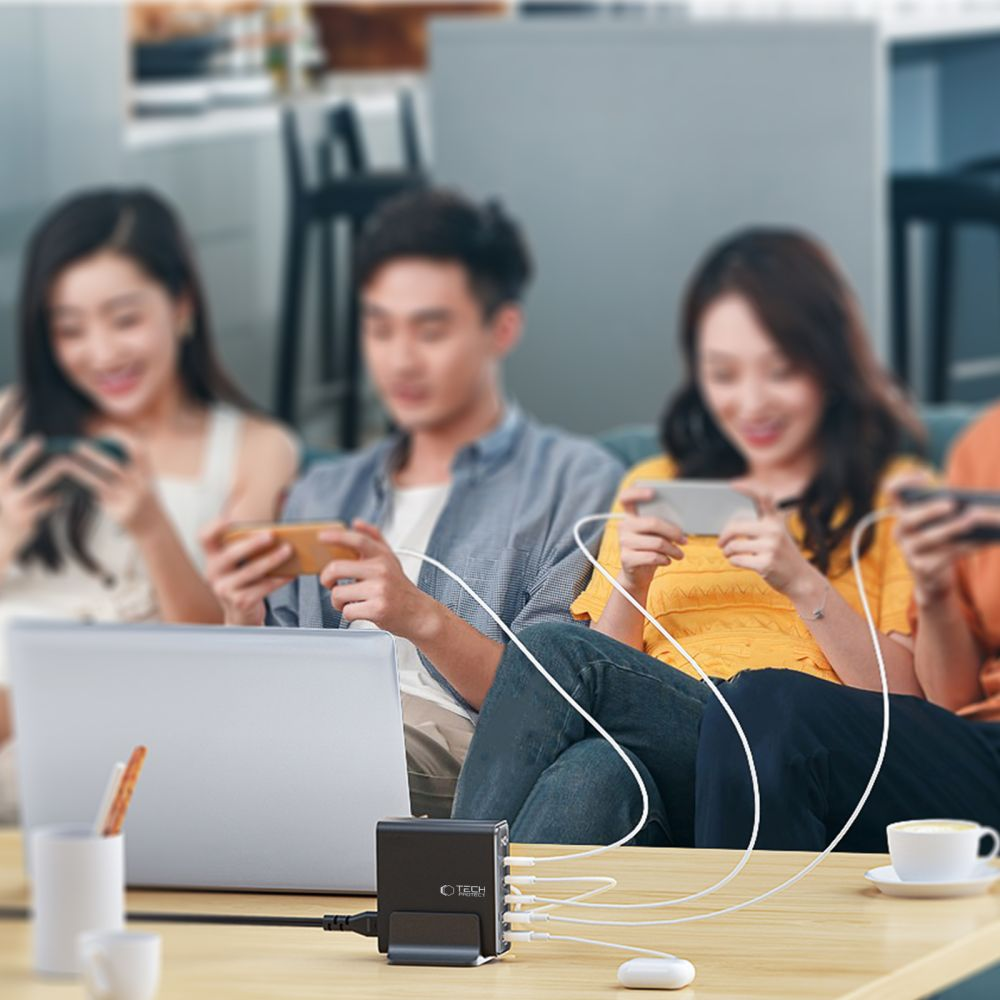 Quatre personnes sur un canapé utilisent des smartphones connectés à un chargeur noir sur une table. Un ordinateur portable et du café sont présents.
