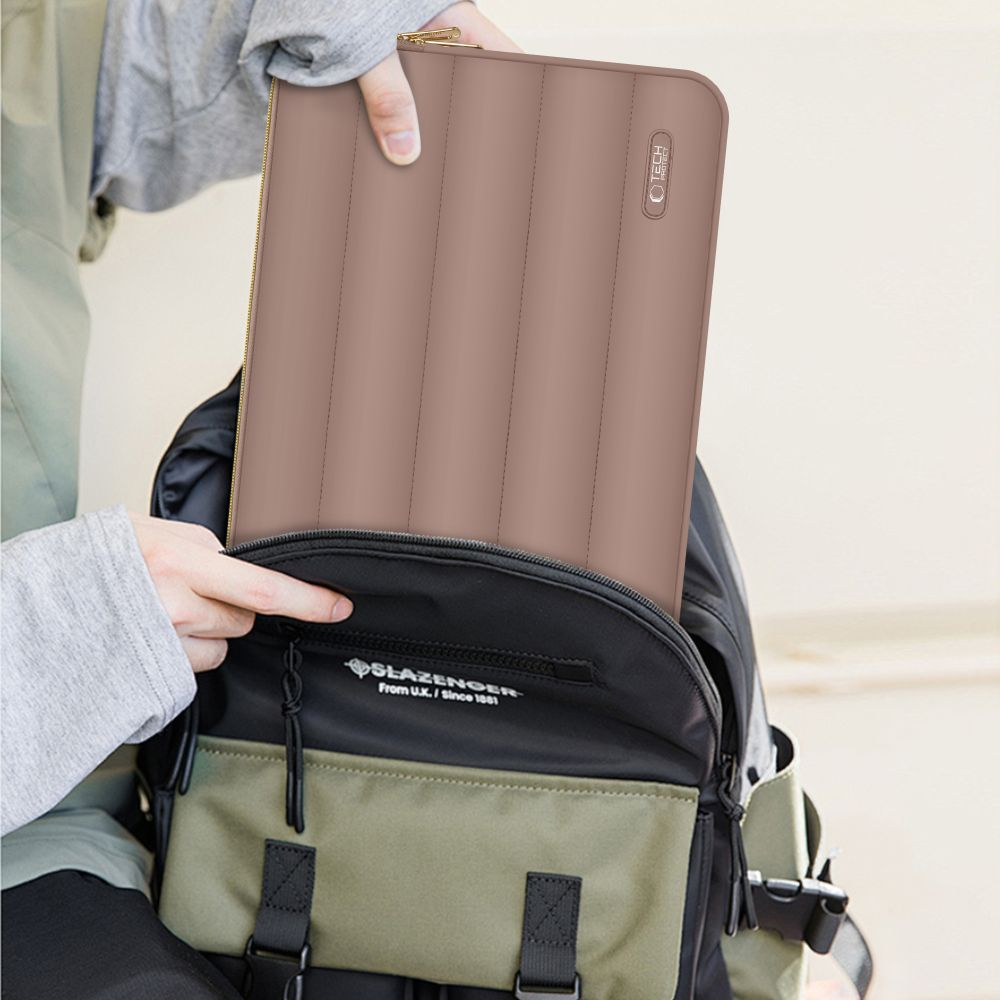 Een persoon plaatst een bruine, gewatteerde laptoptas in een zwarte en groene rugzak. De tas heeft een gouden rits.