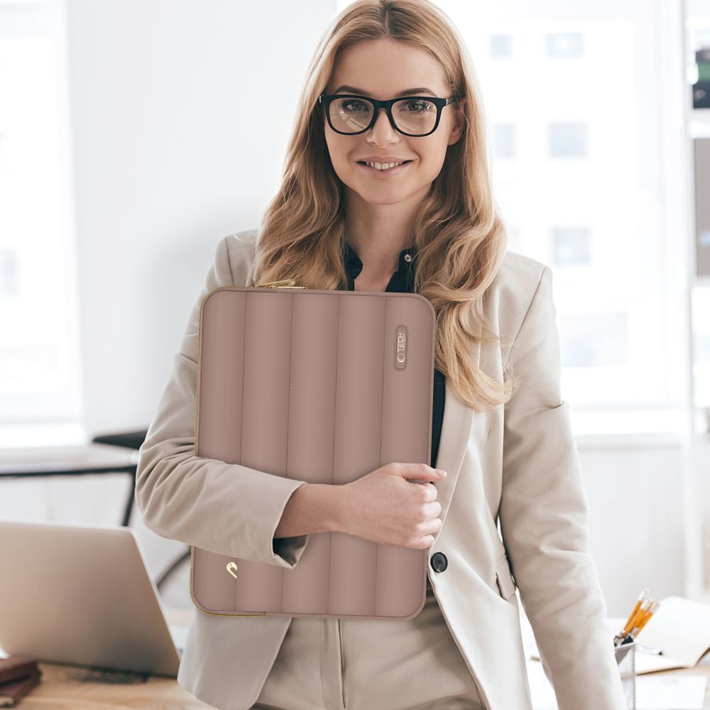 Een vrouw met een bril houdt een roze laptoptas vast. Ze draagt een blazer en glimlacht. Een laptop en kantoorbenodigdheden staan op de achtergrond.