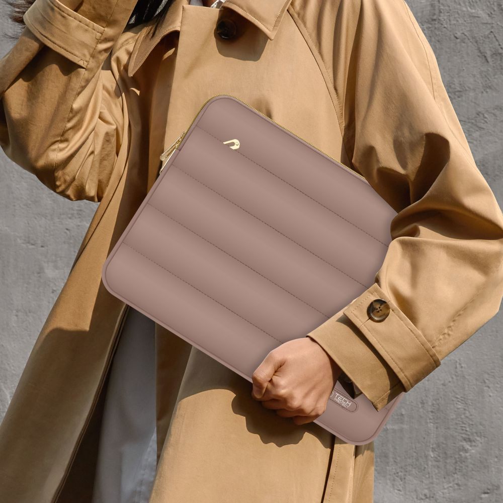 Persoon houdt een mauve laptophoes met gouden rits vast. Ze dragen een beige trenchcoat, tegen een grijze muur.
