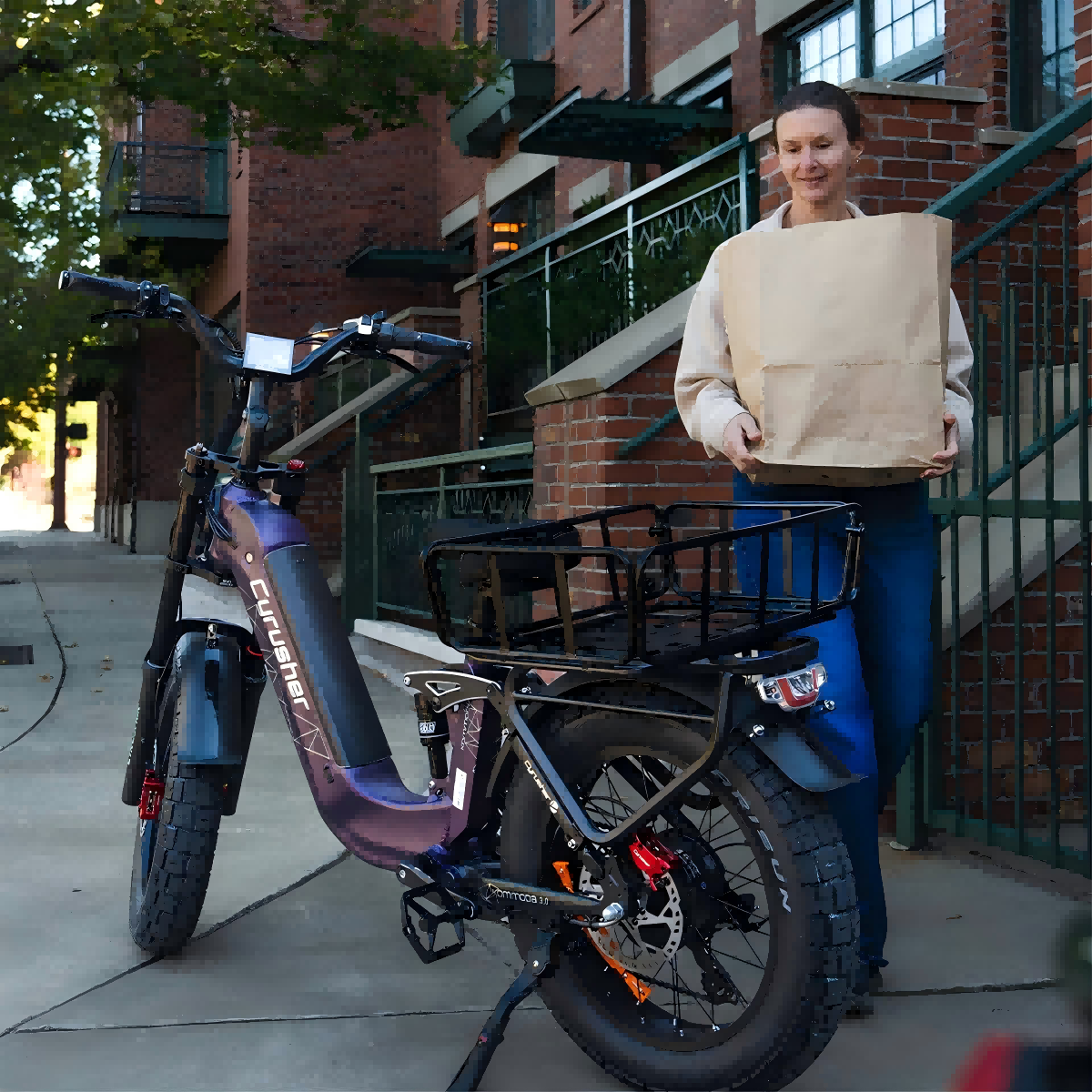Kobieta trzymająca brązową papierową torbę, stojąca obok fioletowego roweru elektrycznego. Ceglany budynek i zielone metalowe ogrodzenie w tle.