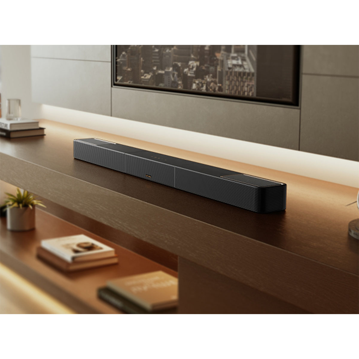 Czarny soundbar stoi na drewnianej półce, przed telewizorem. Na półce są też książki i rośliny, a w tle widać miasto.