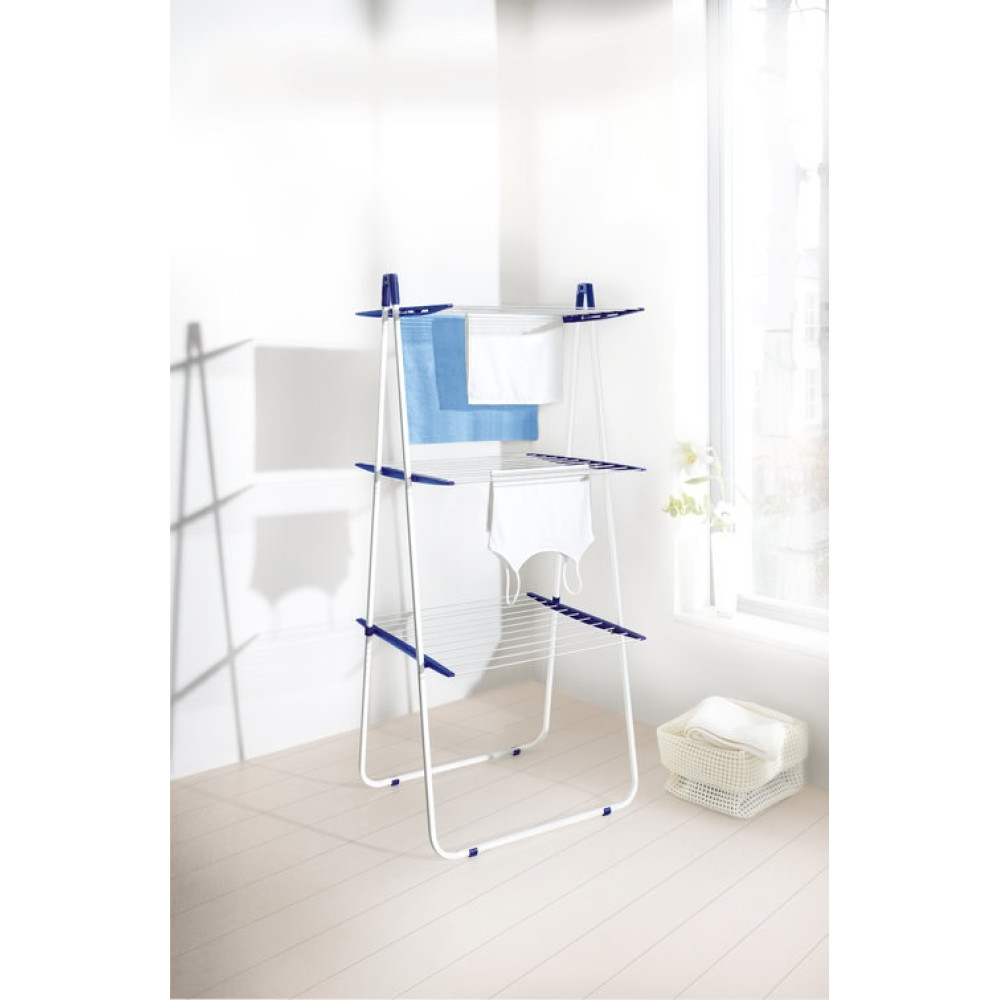 Biało-niebieska suszarka do ubrań z wiszącym praniem. W tle okno ze światłem i kosz ze złożonymi ubraniami.