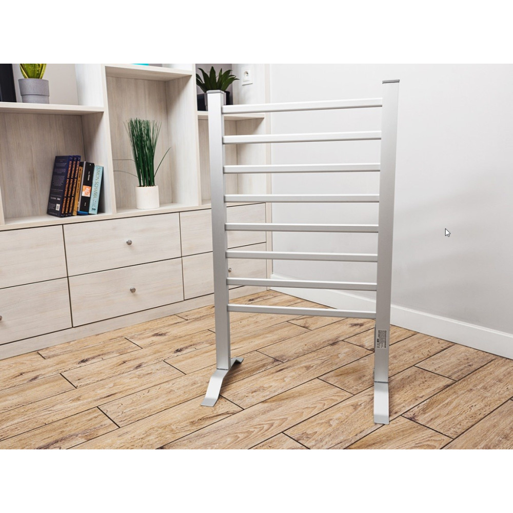 Srebrny podgrzewany wieszak na ręczniki stoi na drewnianej podłodze. Po lewej stronie biała szafka z szufladami i książkami.