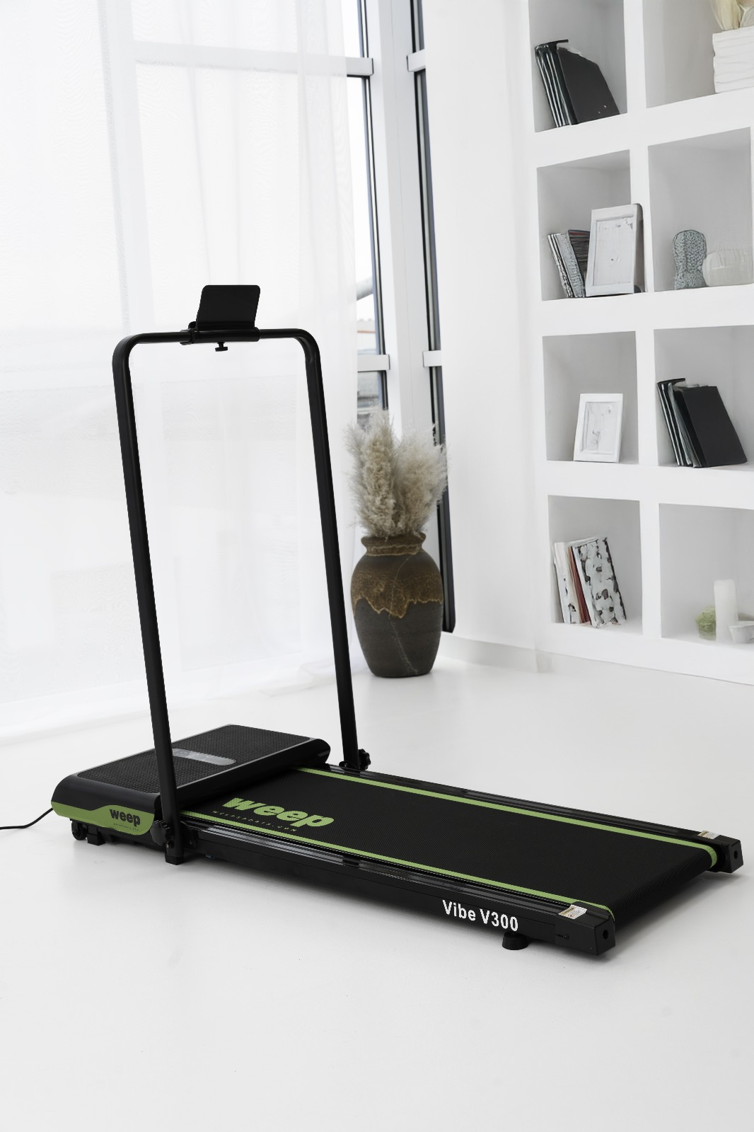 Czarna bieżnia z zielonymi akcentami w białym pokoju. Wazon z suszonymi roślinami i półki z przedmiotami w tle.