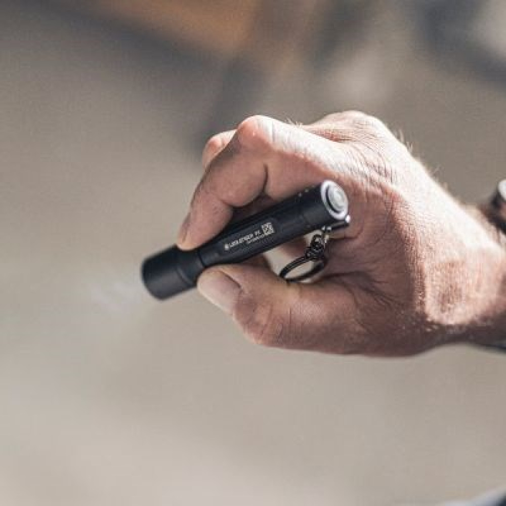 Dłoń trzyma czarną latarkę. Tło jest rozmyte, sugerując otoczenie na zewnątrz. Latarka ma brelok.