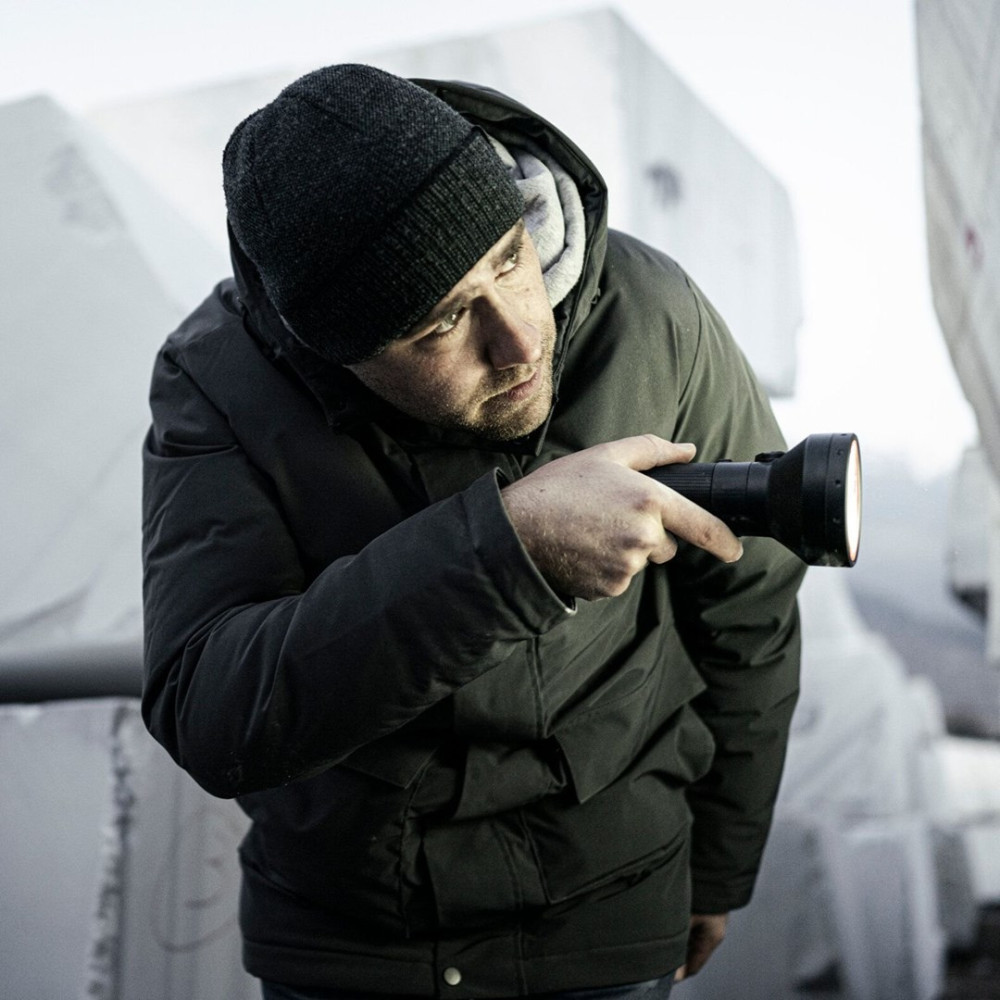 Mężczyzna w czapce i kurtce trzymający latarkę, oświetlający scenę. Tło przedstawia duże, jasne bloki.