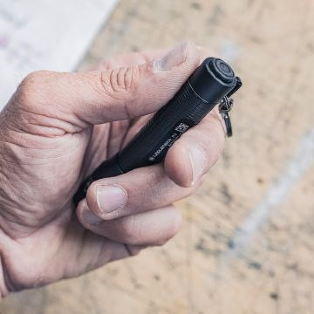 Dłoń trzyma czarną latarkę. Latarka ma kółko na klucze. Tło pokazuje drewnianą powierzchnię i papier.