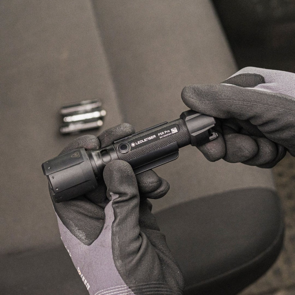 Ręce w rękawicach trzymające czarną latarkę LEDLENSER P5R Pro. W tle wnętrze samochodu z metalowym przedmiotem.