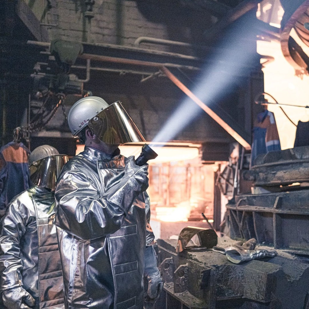 Dwóch pracowników w srebrnych kombinezonach ochronnych w fabryce. Jeden trzyma latarkę. Jasne światło z góry. Tło z maszynami.