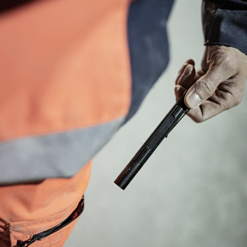 Osoba trzyma czarny długopis. Osoba ma na sobie pomarańczową kamizelkę roboczą. Tło jest jasnoszare.