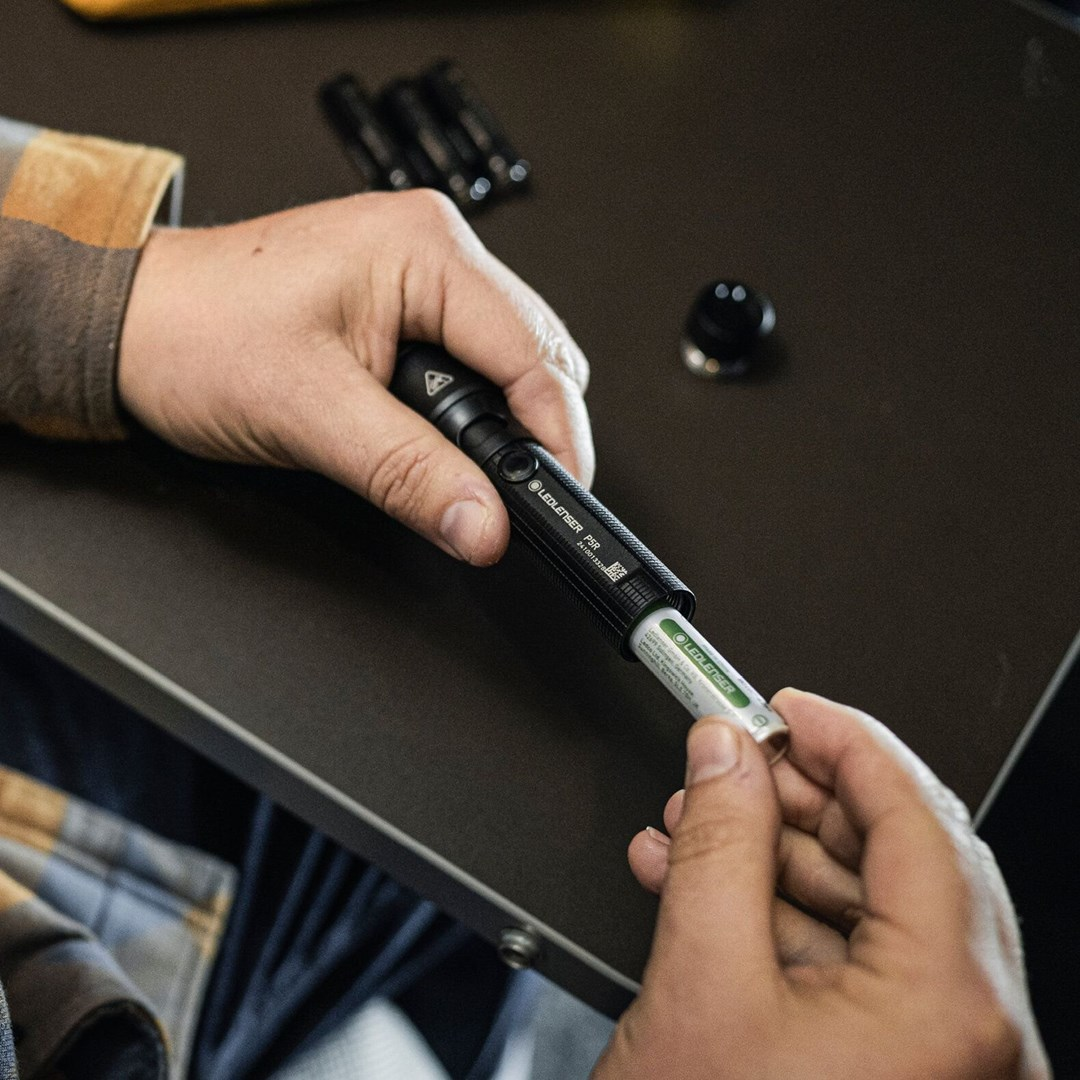 Ręce wkładające baterię do czarnej latarki na ciemnej powierzchni. Inne latarki i czarny obiekt w tle.