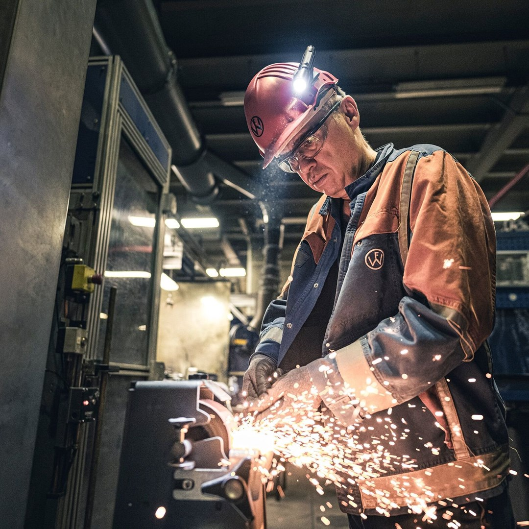 Robotnik szlifuje metal, nosząc kask z lampą. Iskry lecą. Ma na sobie niebieską kurtkę z pomarańczową kamizelką i okulary ochronne.