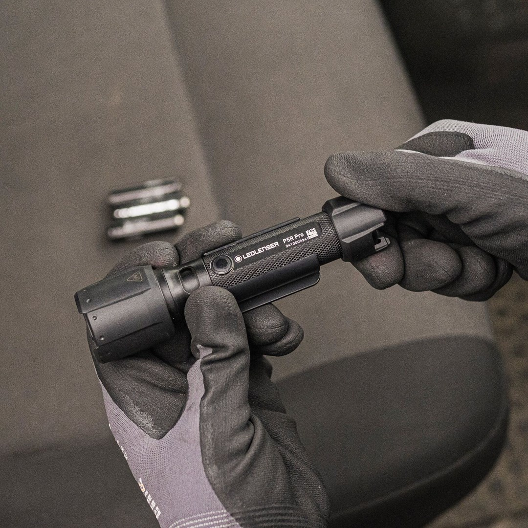Ręce w rękawicach trzymają czarną latarkę Ledlenser P5R Pro. Metalowy przedmiot w tle.