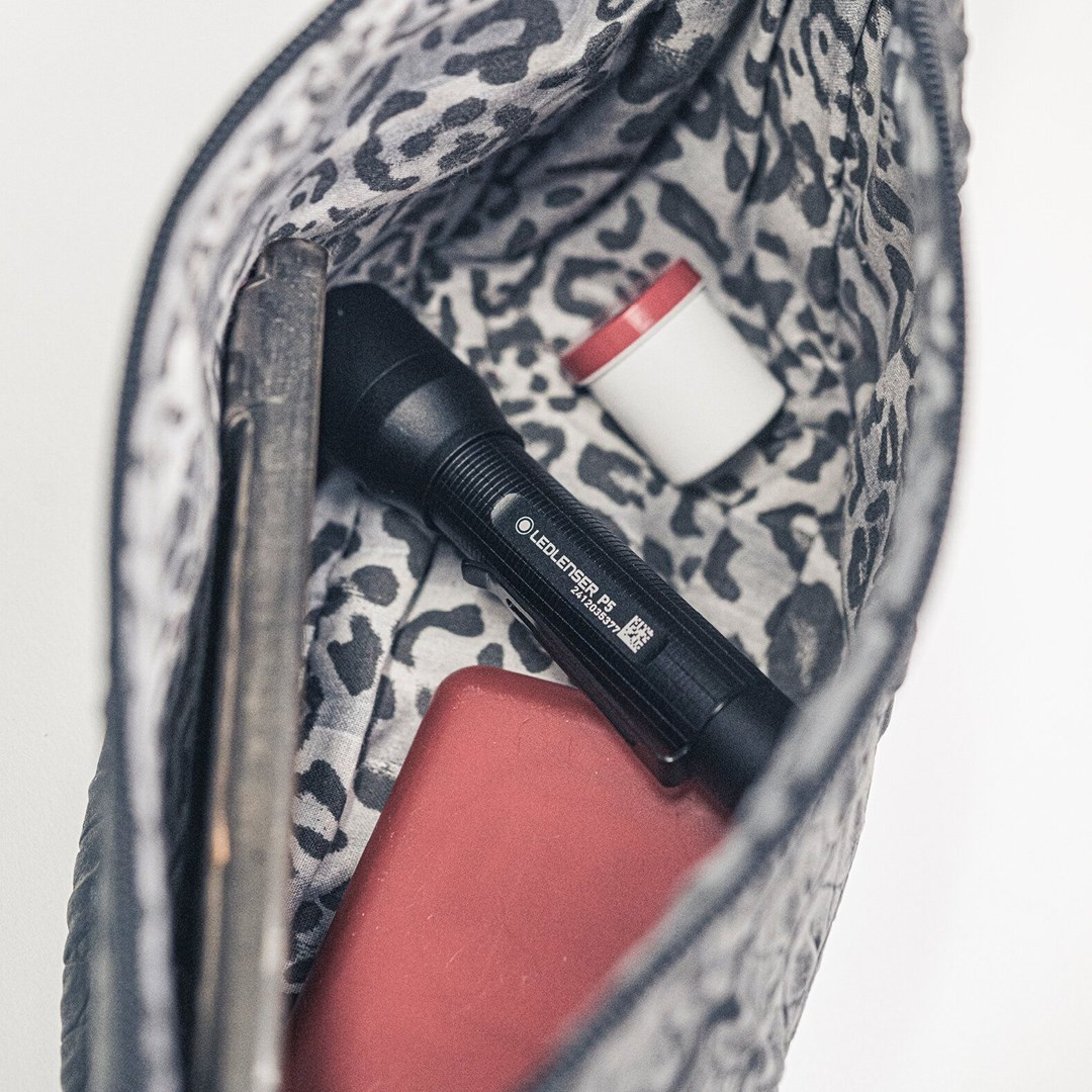 W torbie: czarna latarka, czerwony telefon, biały pojemnik z czerwoną pokrywką na materiale w panterkę. Czarna torba z teksturą.