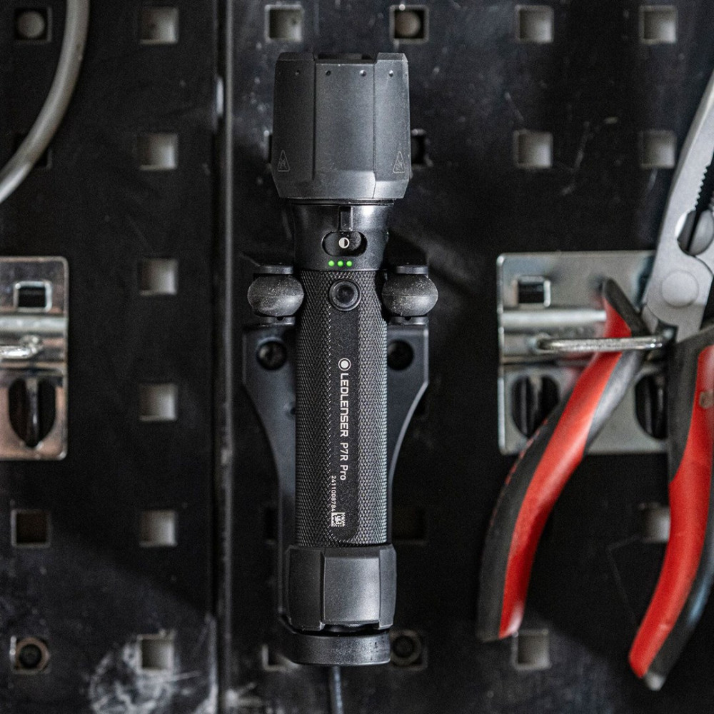 Czarna latarka zamontowana na tablicy perforowanej, szczypce po prawej. Tablica perforowana i narzędzia są ostre. Latarka ma zielone światła.